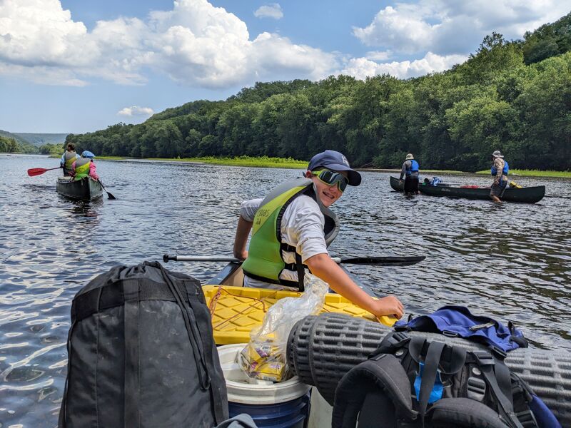 A group of people are canoeing on a river. The person in the foreground is wearing a hat and life vest and is paddling the canoe. There are backpacks and other gear in the canoe. In the background, there are other canoes and trees.