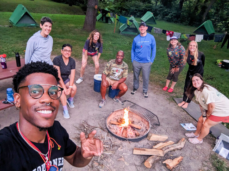 A group of people are gathered around a campfire near several tents. Some individuals are standing and smiling, while others are seated. The atmosphere appears to be one of camaraderie and outdoor enjoyment. The tents suggest they are camping, and the campfire indicates they might be cooking or simply enjoying the warmth and light.