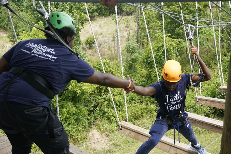 Two people are on a ropes course. One person, wearing a blue shirt, is assisting the other, who is wearing a yellow helmet and a shirt that says "Faith Over Fear", as they navigate a section of the course with wooden planks. They are surrounded by trees and greenery.