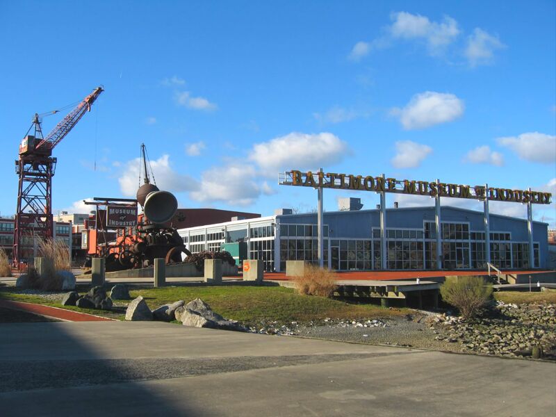 The image shows the Baltimore Museum of Industry. In the foreground, there's a grassy area with some rocks. To the left, there is a large red crane. In the background, there is a blue building with the museum's sign. The sky is blue with some clouds.