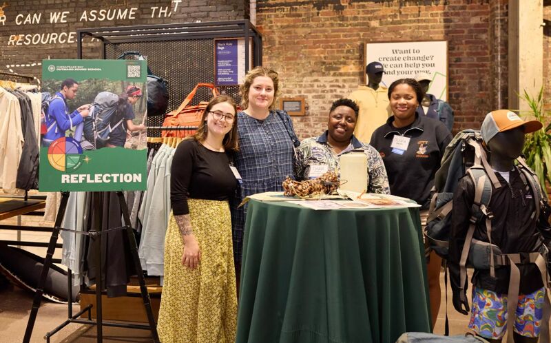 The image shows four women standing near a table with a green tablecloth. There is a sign that says "Reflection". There are also mannequins with backpacks and clothing racks in the background. The setting appears to be an event or a store.