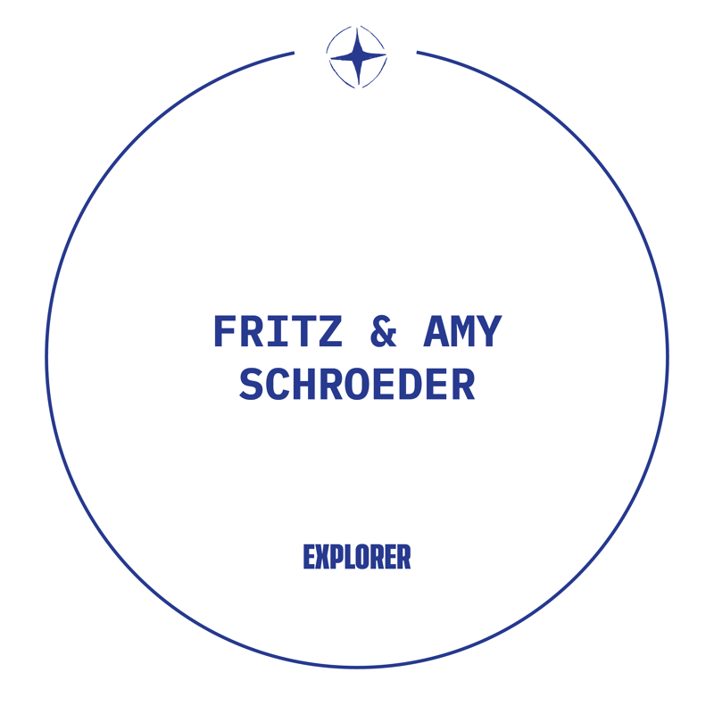 This image features a circular design with the names "FRITZ & AMY SCHROEDER" prominently displayed in the center in a bold, dark blue font. Below their names, the word "EXPLORER" is also written in the same font. A small, stylized compass rose graphic is positioned at the top center, just inside the circle's border. The overall aesthetic is clean and modern, suggesting a personal brand or a commemorative item related to exploration.