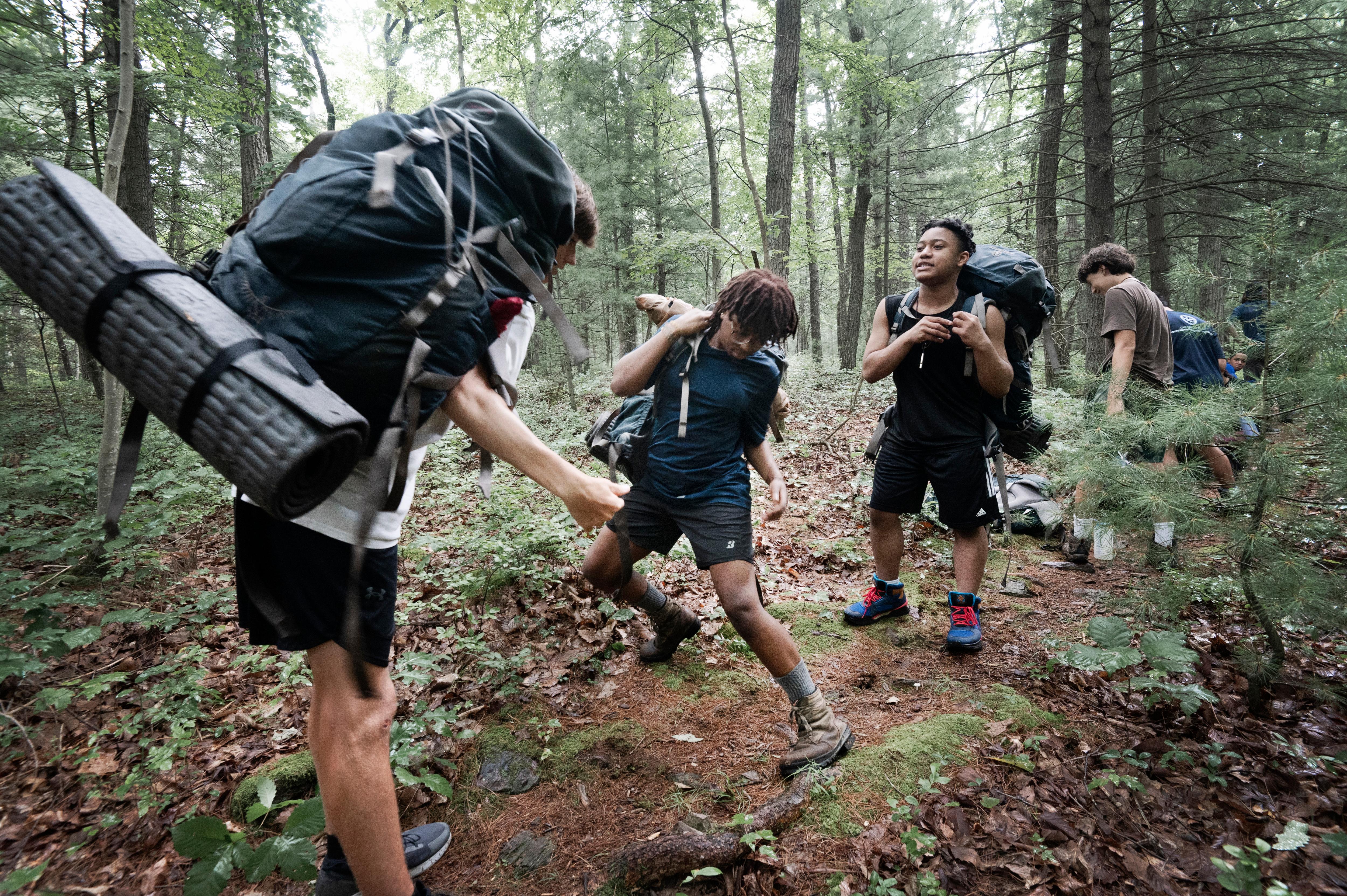 The image shows a group of young people hiking in a forest. They are all carrying backpacks, and some have sleeping mats attached. The hikers are walking on a dirt path through the trees. The forest is dense and green, with tall trees and thick undergrowth. The hikers appear to be enjoying their time in nature.