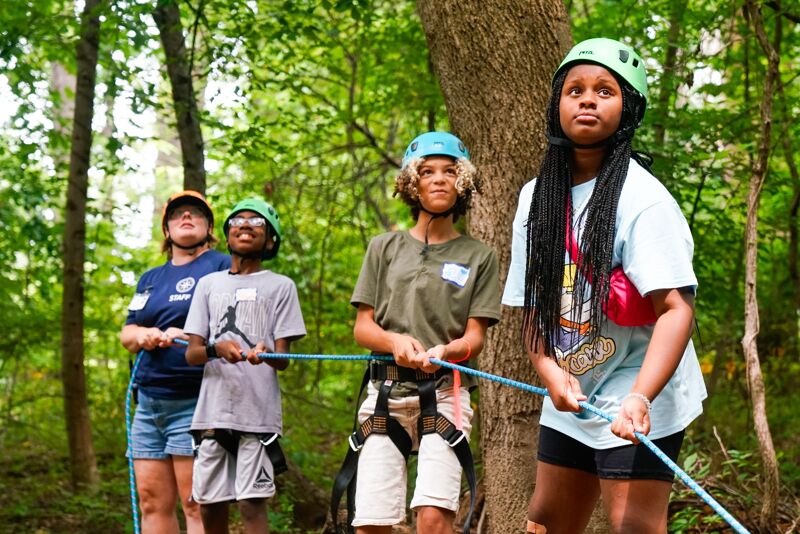 The image shows four young people in a wooded area, likely participating in an outdoor activity or ropes course. They are all wearing helmets and appear to be holding onto ropes or cables. The focus is on the girl in the front, who is looking upwards with a determined expression. The background is filled with trees and foliage, suggesting a natural setting.