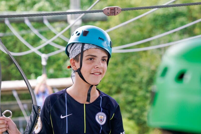 A young boy wearing a blue helmet and a navy blue t-shirt is participating in a ropes course. He is looking to the right with a slight smile. The background shows trees and other parts of the ropes course, including wires and support structures. Another person wearing a green helmet is visible in the lower right corner of the image.