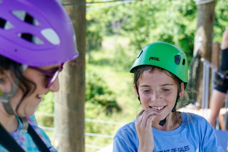 The image shows two people wearing helmets, likely participating in an outdoor activity. The person on the left has a purple helmet and is partially visible. The person on the right has a green helmet and is looking to the left with a slight smile. They appear to be in a wooded area, possibly an adventure park or ropes course.