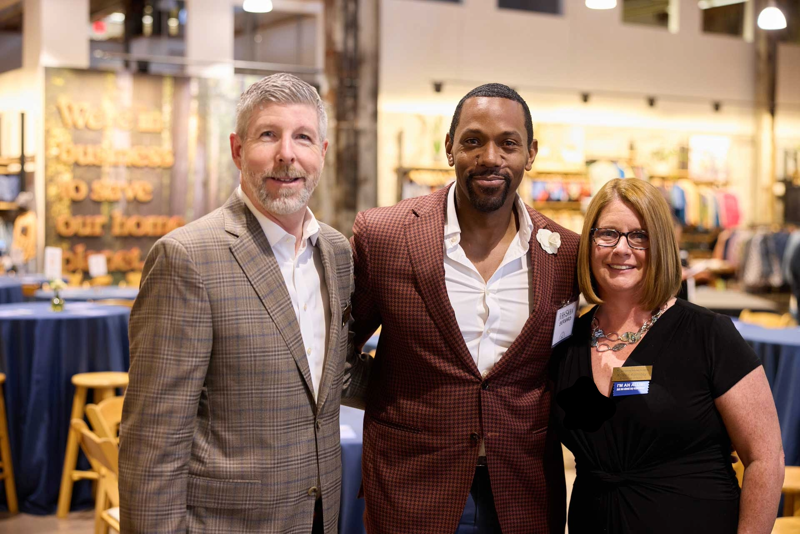 The image shows three people posing indoors. A man in a brown checkered blazer stands on the left. In the center is a man in a maroon suit jacket and white shirt. A woman in a black dress stands on the right. They are all smiling and appear to be at an event or gathering.
