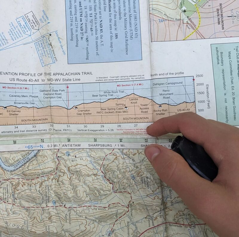 A person's hand is holding a black object over a map. The map features an elevation profile of the Appalachian Trail, specifically from US Route 40-Alt to the MD-WV State Line. Text on the map includes place names like "Antietam" and "Sharpsburg," along with elevation markers and other geographical details. The map appears to be a topographical or hiking map.