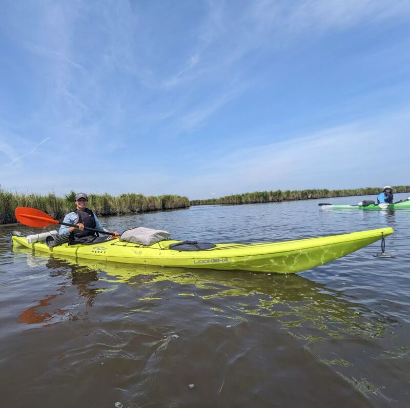 A person is kayaking on a sunny day in a bright yellow kayak. They are holding an orange paddle and wearing a hat and life vest. The water is calm, and there is green vegetation on the shoreline. The sky is blue with some clouds. Another person is visible in the distance, also on the water.