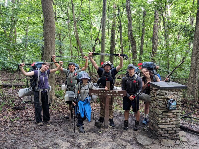 A group of seven people are standing in a forest, posing for a photo. They are all wearing backpacks and hiking gear, suggesting they are on a hiking trip. The group is gathered around a stone pillar with a wooden sign on top. The forest is lush and green, with tall trees surrounding the group.