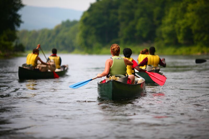 The image shows a group of people canoeing down a river. There are two canoes visible, each carrying several people. The people in the canoes are paddling, and the canoes are moving through the water. The river is surrounded by trees, and the sky is overcast. The overall scene suggests a recreational or team-building activity in a natural setting.