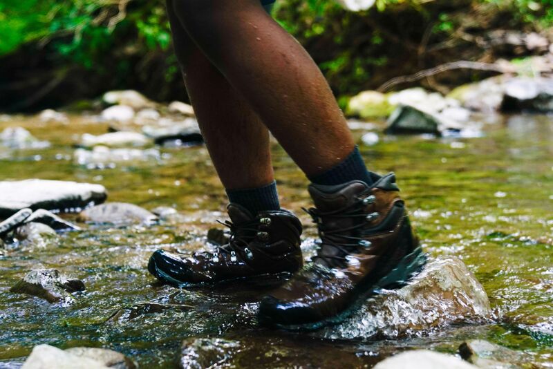 The image shows a person wearing hiking boots standing in a shallow stream. The water is flowing around the rocks and the person's boots. The person is wearing dark socks and their legs are visible. The hiking boots appear to be waterproof, as the water is beading up on the surface. The background consists of rocks and foliage, suggesting a natural outdoor setting. The lighting is bright, indicating it is daytime.