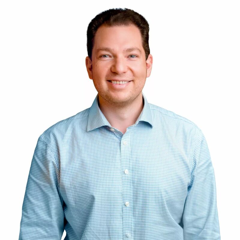 The image shows a man with fair skin and short, dark hair. He is wearing a light blue, button-down shirt with a subtle checkered pattern. The man is smiling, and the background is plain white. The overall impression is clean and professional.