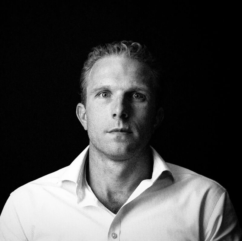 The image shows a man in a white shirt against a black background. He has short, light-colored hair and is looking directly at the camera with a neutral expression. The lighting is focused on his face, highlighting his features. The overall impression is one of simplicity and directness.