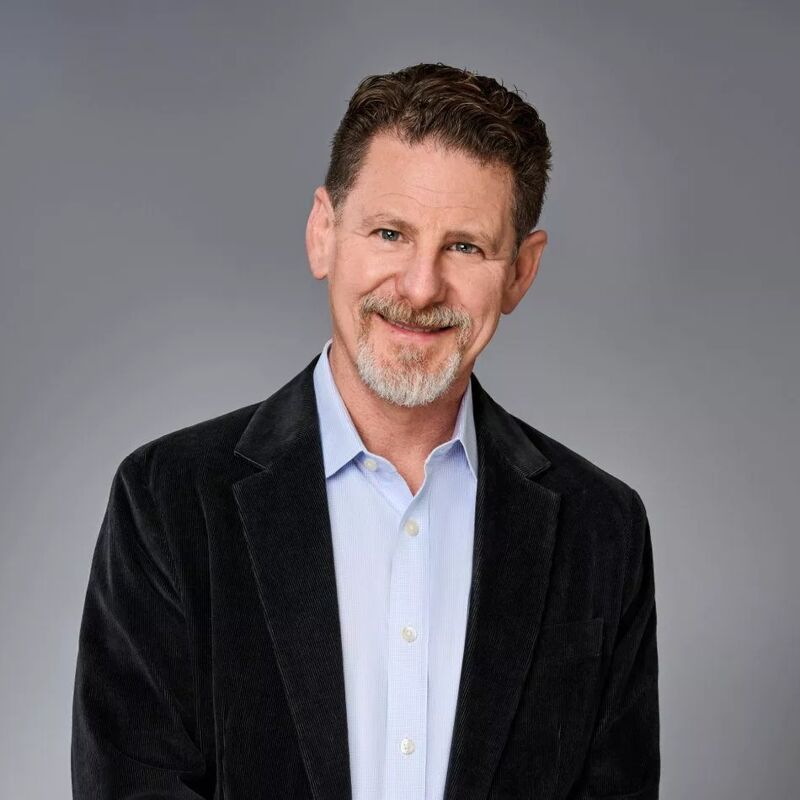 The image shows a middle-aged man with short, graying hair and a neatly trimmed beard. He is wearing a light blue shirt under a dark jacket. He is smiling slightly, and the background is a plain gray. The man appears to be in a studio setting.