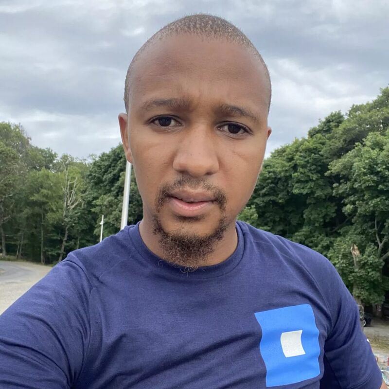 The image shows a close-up of a man with short, dark hair and a goatee. He is wearing a blue t-shirt with a white and blue square design on the chest. The background includes trees and a cloudy sky. The man is looking directly at the camera.