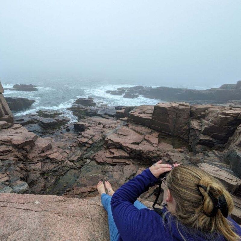 The image shows a woman sitting on a rocky cliff overlooking the ocean on a foggy day. She is holding a camera, suggesting she is taking a picture of the scenery. The rocks are reddish-brown, and the water is choppy. The overall atmosphere is serene and a bit mysterious due to the fog.