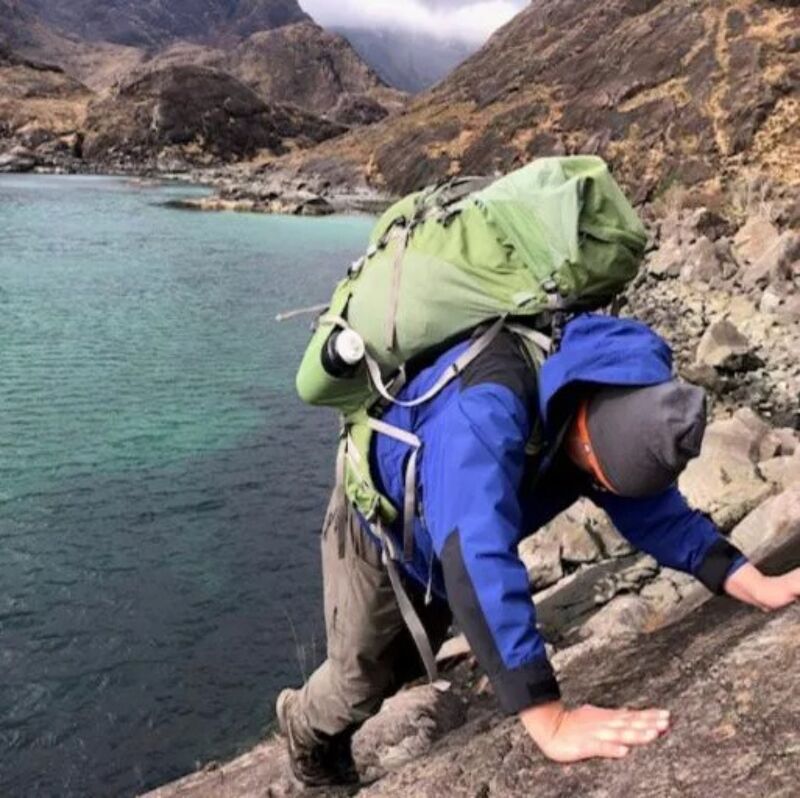 The image shows a person hiking near a body of water. The person is wearing a blue jacket, a backpack, and is climbing on rocks. The water is a beautiful turquoise color, and the background features mountains. The scene suggests an adventurous outdoor activity.