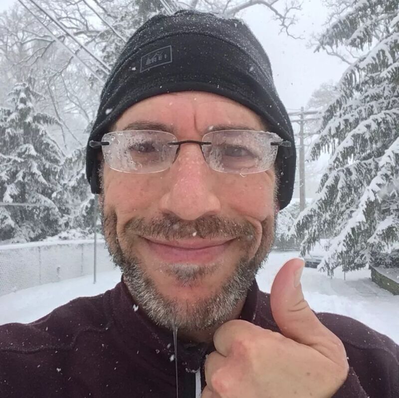 The image shows a man giving a thumbs up in a snowy environment. He is wearing a black beanie and glasses covered in ice. The background features snow-covered trees and a fence. The man appears to be enjoying the cold weather.