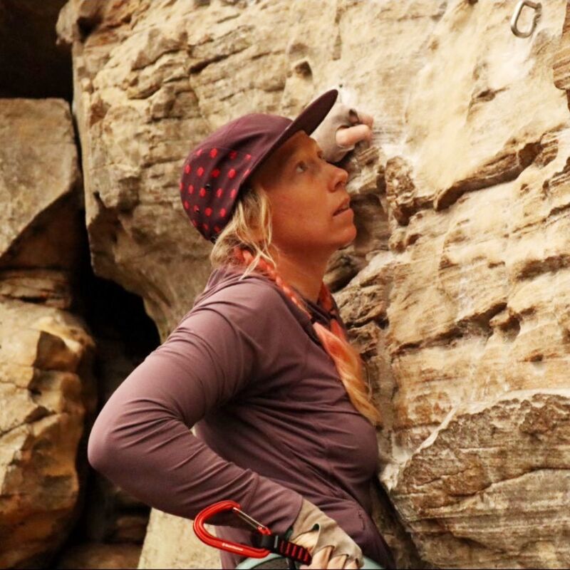 The image shows a woman rock climbing. She is wearing a cap, long-sleeved shirt, and gloves. She is looking up at the rock face, with one hand gripping a hold. A carabiner is attached to her harness. The rock appears to be sandstone.