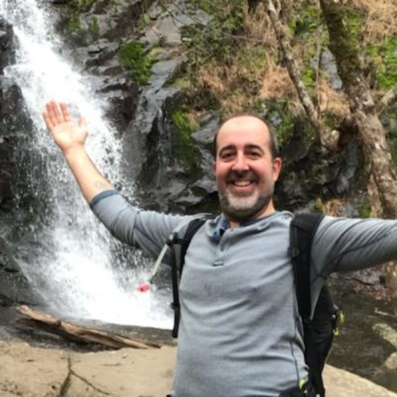 A man with a beard and a backpack stands in front of a waterfall with his arms outstretched. He is wearing a gray long-sleeved shirt and appears to be smiling. The waterfall cascades down rocks in the background, surrounded by greenery. The man seems to be enjoying the scenery and embracing the natural environment.