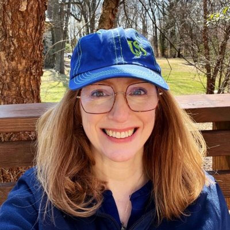 The image shows a woman with red hair smiling at the camera. She is wearing glasses and a blue baseball cap with a green logo. She appears to be sitting outside on a wooden deck or porch, with trees visible in the background. The lighting suggests it is daytime.