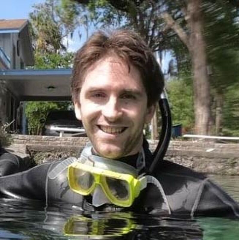 The image shows a man in a wetsuit and snorkeling gear, partially submerged in water. He is smiling and appears to be looking directly at the camera. The background includes trees and a building, suggesting an outdoor location, possibly a lake or river. The lighting is bright, indicating it is daytime.