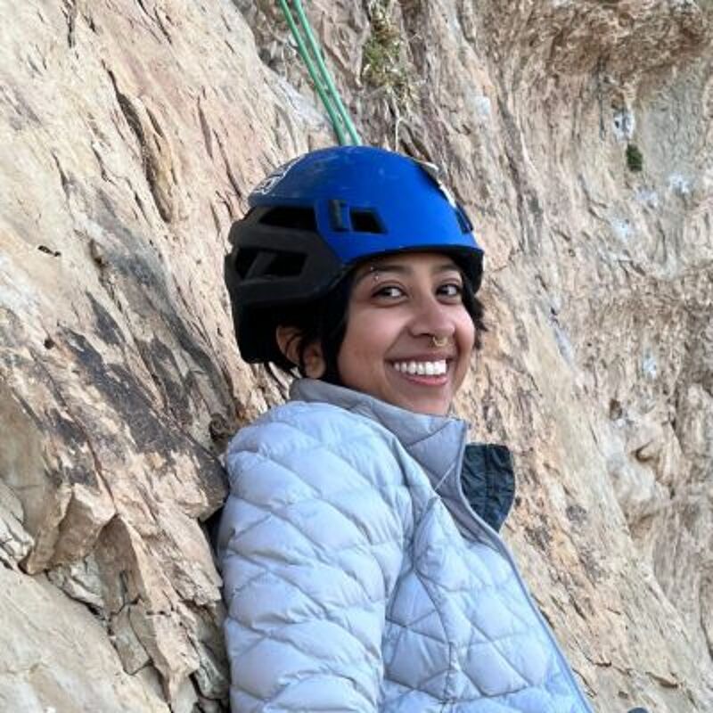 A woman with a blue climbing helmet smiles at the camera. She is wearing a light gray jacket. Behind her is a rock wall, suggesting she is rock climbing. A green rope is visible above her, further indicating her activity. The lighting is bright, highlighting the textures of the rock and her gear.