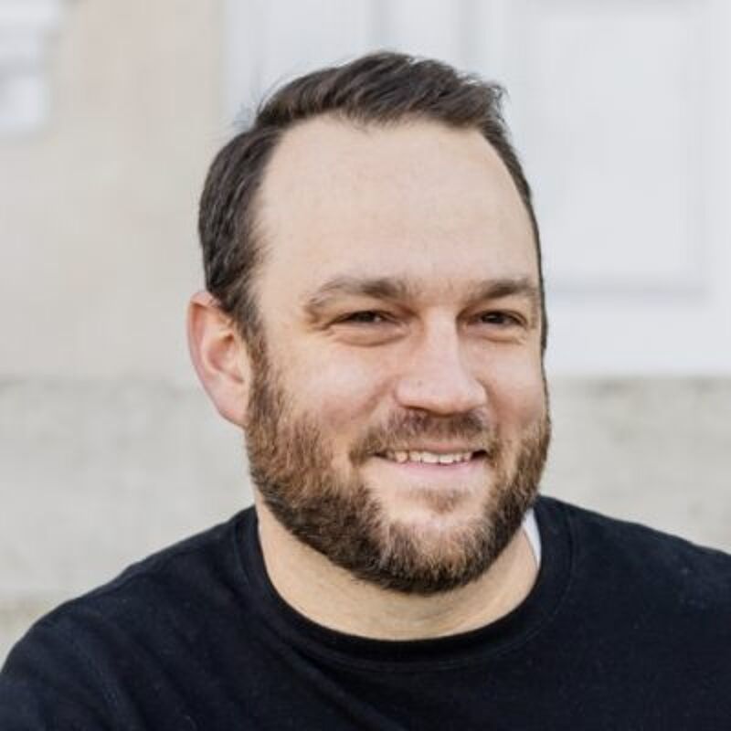 The image shows a man with a beard and short brown hair. He is wearing a black shirt and has a light skin tone. The background appears to be a building with white trim and steps. The man is smiling slightly and looking directly at the camera. The photo is well-lit and in focus.