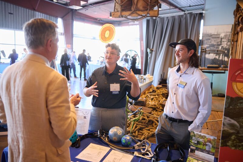 In the image, three people are engaged in a conversation at what appears to be an event or exhibition. The person in the center, wearing a dark shirt, is actively gesturing while speaking to the other two individuals. The person on the right, in a light-colored shirt and baseball cap, is smiling and listening. The person on the left, seen from the back, is also facing the speaker. Various items, including globes and ropes, are visible on a table in the foreground.