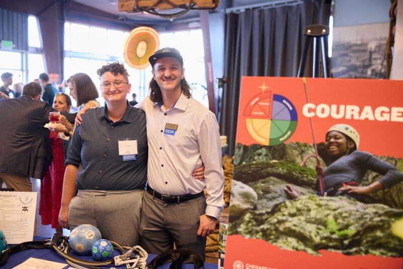Two individuals, one wearing glasses and a dark blue collared shirt, and the other with a mustache and a light-colored collared shirt, pose for the camera. They are standing in front of a table with various items, including globes and what appear to be climbing ropes. Behind them, a large sign features the word "COURAGE" and an image of someone rock climbing. The overall setting seems to be an indoor event or gathering.