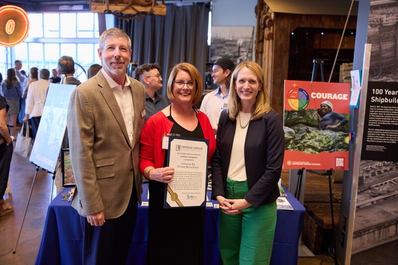In the image, three people are posing for a photo. On the left is a man with a beard and a plaid jacket. In the middle is a woman in a red cardigan holding a certificate. On the right is a woman in a dark blazer and green pants. They are standing in front of displays, possibly at an event or exhibition.