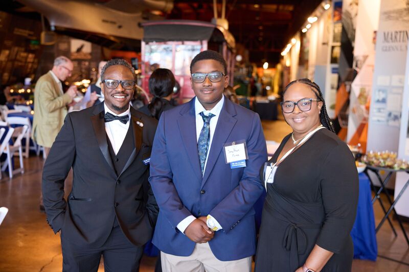 Three people, two men and one woman, are posing for a photo indoors. The man on the left is wearing a black tuxedo with a bow tie. The man in the middle is wearing a blue suit with a tie. The woman on the right is wearing a black dress. All three are wearing glasses and smiling. They appear to be at a formal event.