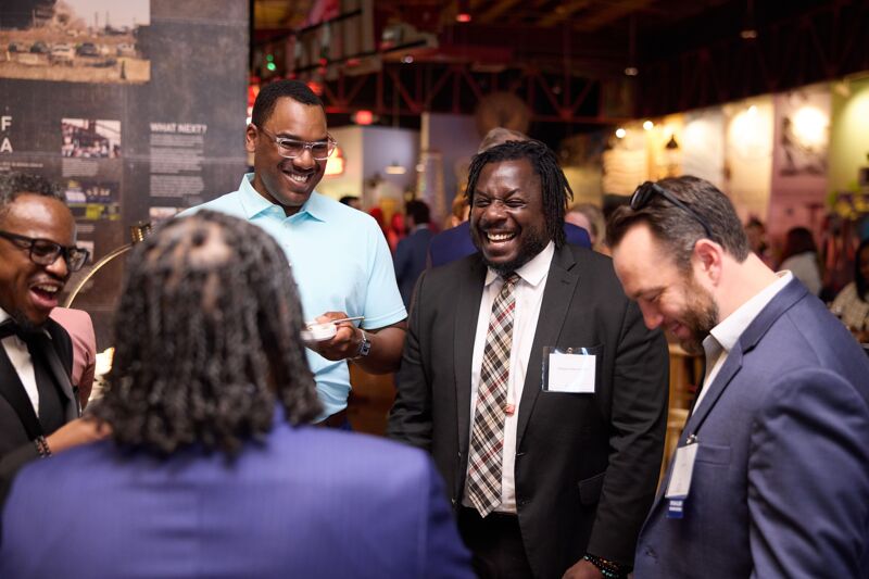 The image shows a group of four men in business attire, engaged in a lively conversation at what appears to be a networking event or a formal gathering. They are all smiling and seem to be enjoying each other's company. The man in the light blue shirt is laughing heartily, as is the man in the black suit next to him. The man on the far right is also smiling, looking down at the man in the foreground. The man on the far left is partially visible, also smiling. The background suggests an indoor venue with other people present.