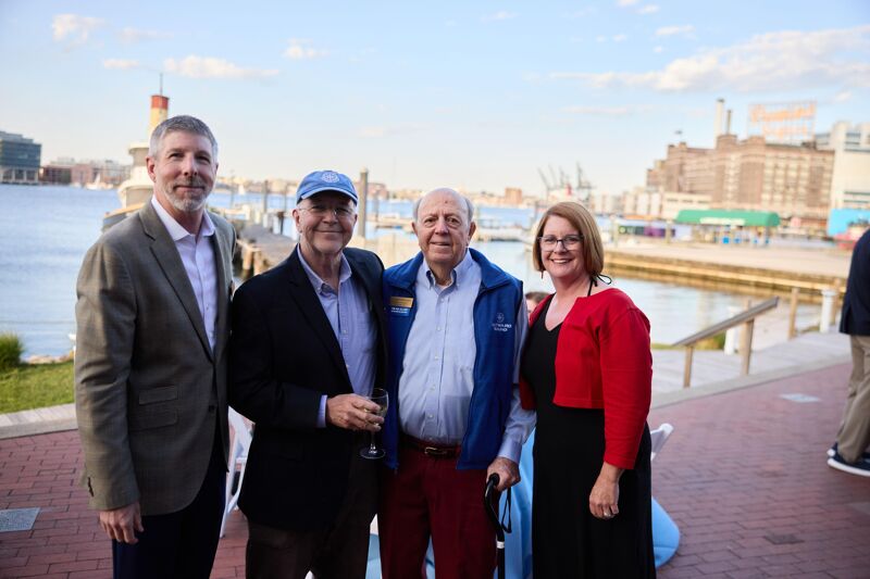 Four people are posing for a photo outdoors with a waterfront in the background. From left to right, a man in a suit, a man in a suit and baseball cap, a man in a blue jacket, and a woman in a red cardigan. The man in the blue jacket is holding a glass. The buildings and water suggest an urban harbor setting.