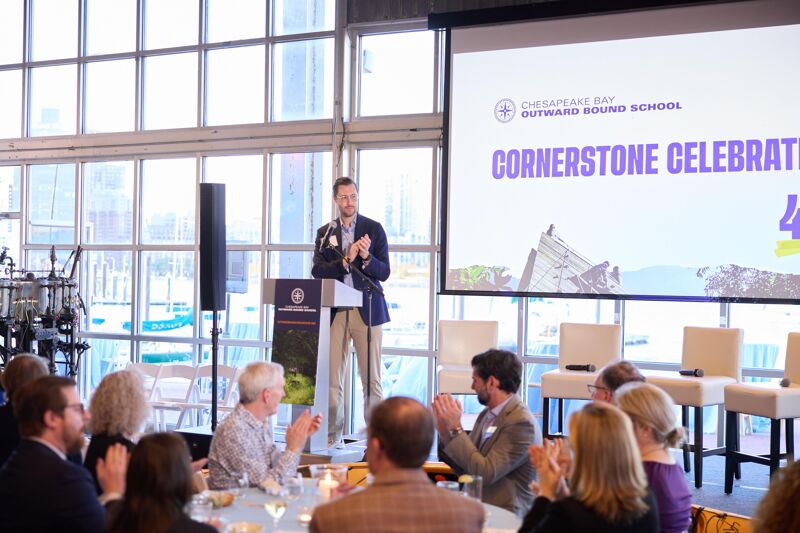 A man in a blue blazer and khaki pants stands at a podium, clapping his hands. He is speaking at an event called "Cornerstone Celebration" for Chesapeake Bay Outward Bound School. The event appears to be a formal gathering with tables set for a meal and an audience seated in chairs. The room has large windows with natural light.