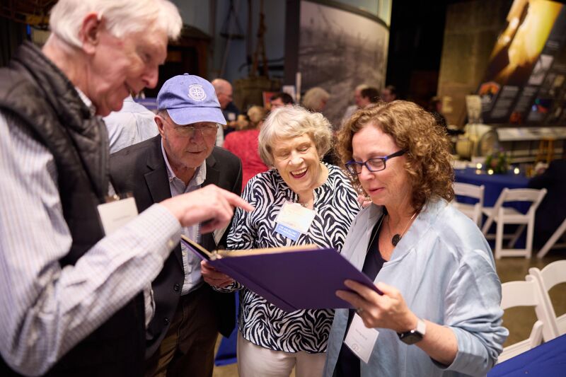 In this image, a group of four older adults are gathered around a table, looking at a purple book. The man on the left is pointing at something in the book, and the others are attentively observing. They appear to be engaged in a lively discussion or sharing of memories. The setting seems to be an indoor event or gathering, possibly a museum or a historical society, with other people and tables visible in the background.
