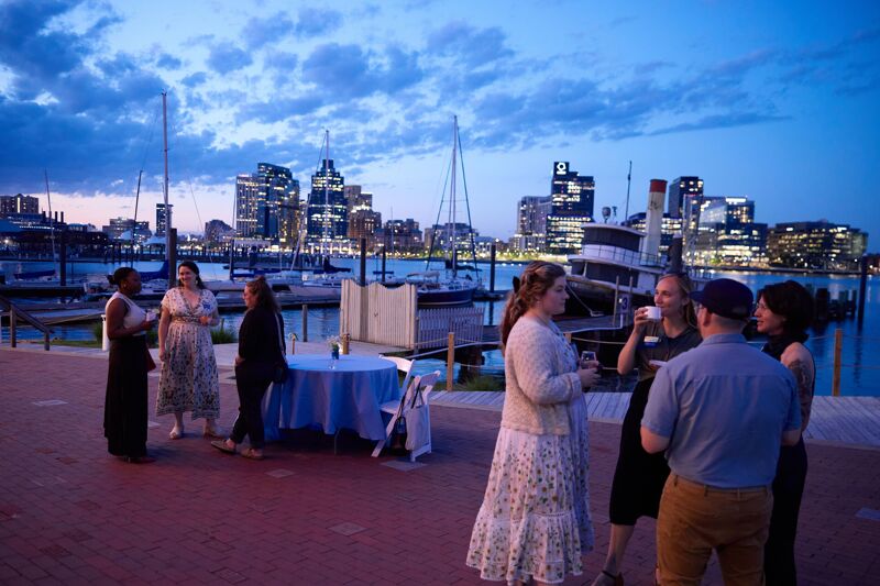 A group of people are gathered on a brick patio near a body of water at dusk. In the background, a city skyline with illuminated buildings is visible, along with several boats docked in a harbor. The sky is a deep blue with hints of orange and purple, suggesting the sun has recently set. Some individuals are dressed formally, while others are in more casual attire, and they appear to be socializing.