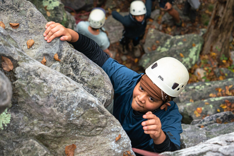 A young person wearing a white helmet is rock climbing. They are gripping the rock face with their hands and looking upwards with a determined expression. In the background, other people, also wearing helmets, are visible among the rocks and trees, suggesting a group activity or climbing class.