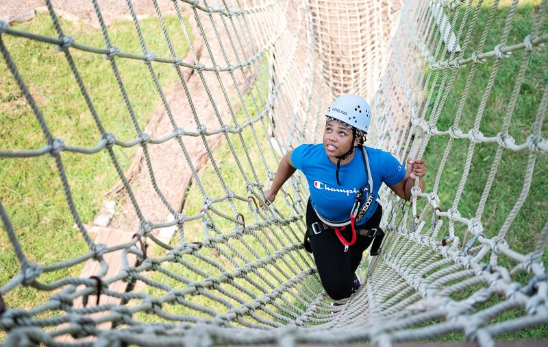 A person wearing a blue t-shirt and a white helmet is climbing a rope bridge. The bridge is made of thick ropes and has a net-like structure. The person is holding onto the ropes for support and appears to be focused on climbing. The background shows green grass and trees, suggesting that the bridge is part of an outdoor adventure course.