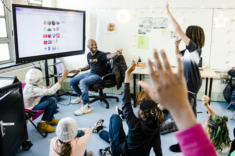 A classroom scene shows a man presenting on a large screen to a group of students. Some students are raising their hands, eager to participate. The atmosphere appears engaging and interactive, suggesting an active learning environment. The room is bright, and the students seem enthusiastic about the lesson.
