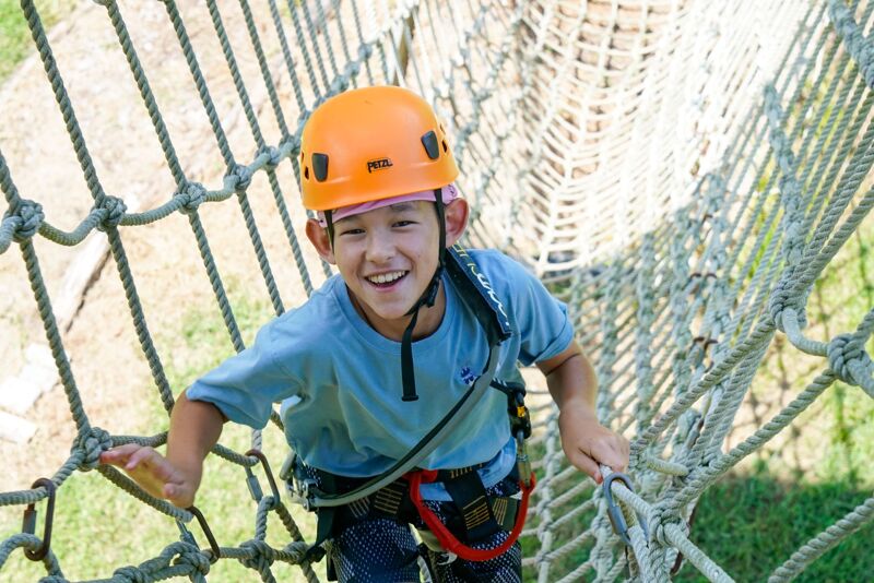 A young boy wearing an orange helmet and a blue t-shirt is smiling while climbing on a rope structure. He appears to be enjoying an outdoor adventure activity, possibly at a ropes course or adventure park. The boy is holding onto the ropes with his hands, and the background shows greenery, suggesting a natural setting.