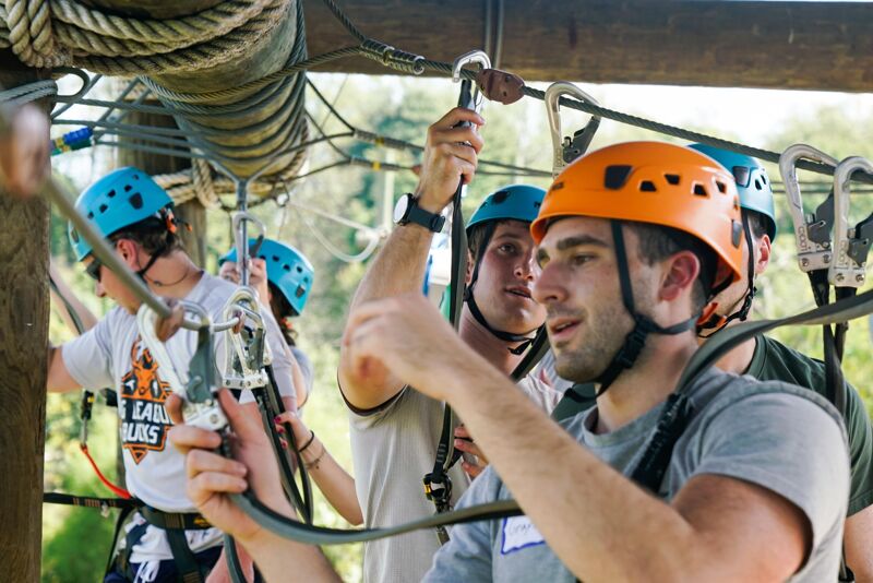 The image shows three people wearing helmets and safety harnesses, likely participating in a ropes course or zip-lining activity. The person in the foreground is holding onto a rope and appears to be adjusting their equipment. The background shows trees and other course elements, suggesting an outdoor adventure setting.
