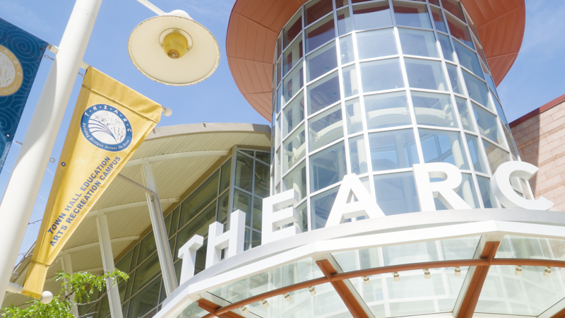 The image shows a modern building with a glass facade and the word "THE ARC" prominently displayed. A banner with a logo and text that reads "TOWN HALL EDUCATION ARTS RECREATION CAMPUS" is visible on the left. The building's architecture features curved lines and a light-colored exterior, suggesting it is a public or community center.