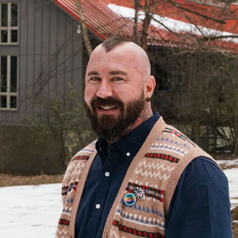 The image shows a man with a shaved head and a mohawk. He has a beard and is wearing a sweater vest over a button-down shirt. He is smiling at the camera. The background includes a building and some snow.