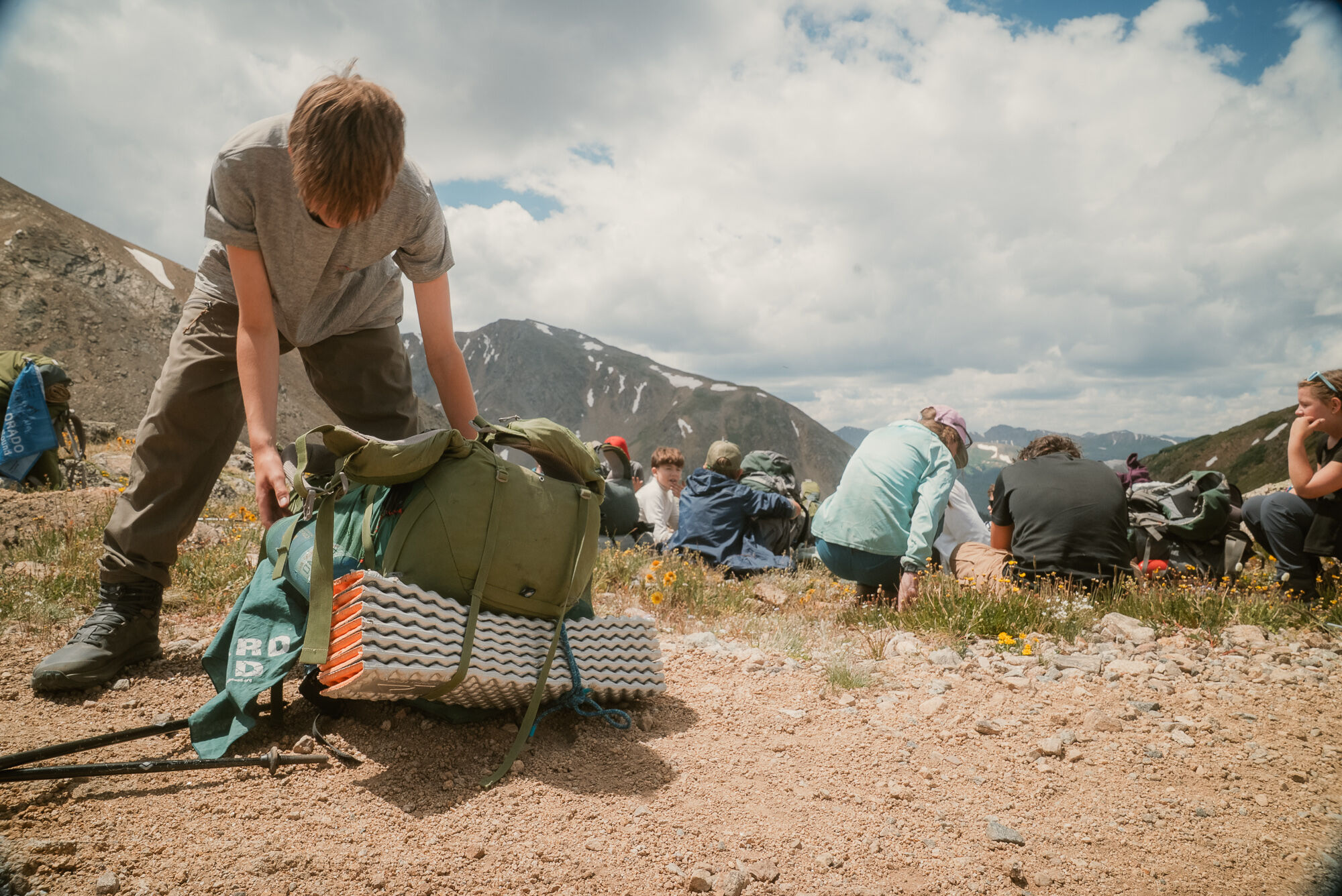 A person is packing a green backpack on a rocky, mountainous terrain. Other people are sitting and resting in the background, enjoying the scenery. The sky is partly cloudy, suggesting a bright day for hiking. The overall setting indicates an outdoor adventure, possibly a group hiking or camping trip in the mountains.
