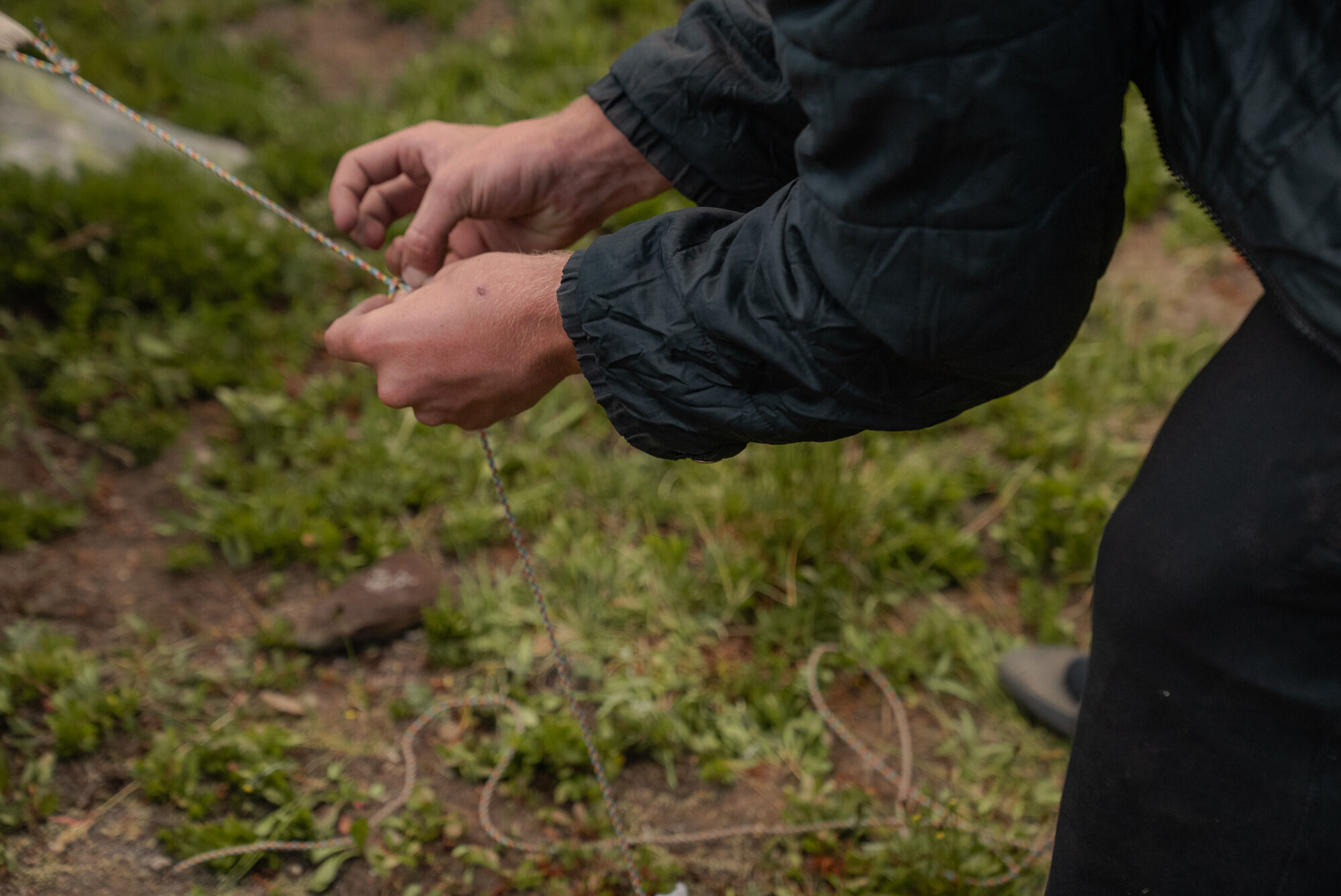 The image shows a close-up of a person's hands as they work with a string or rope. The person is outside, as suggested by the green grass in the background. The hands are the primary focus, with the person's sleeves and part of their body visible. The lighting appears natural, and the overall scene suggests an activity involving manual dexterity.