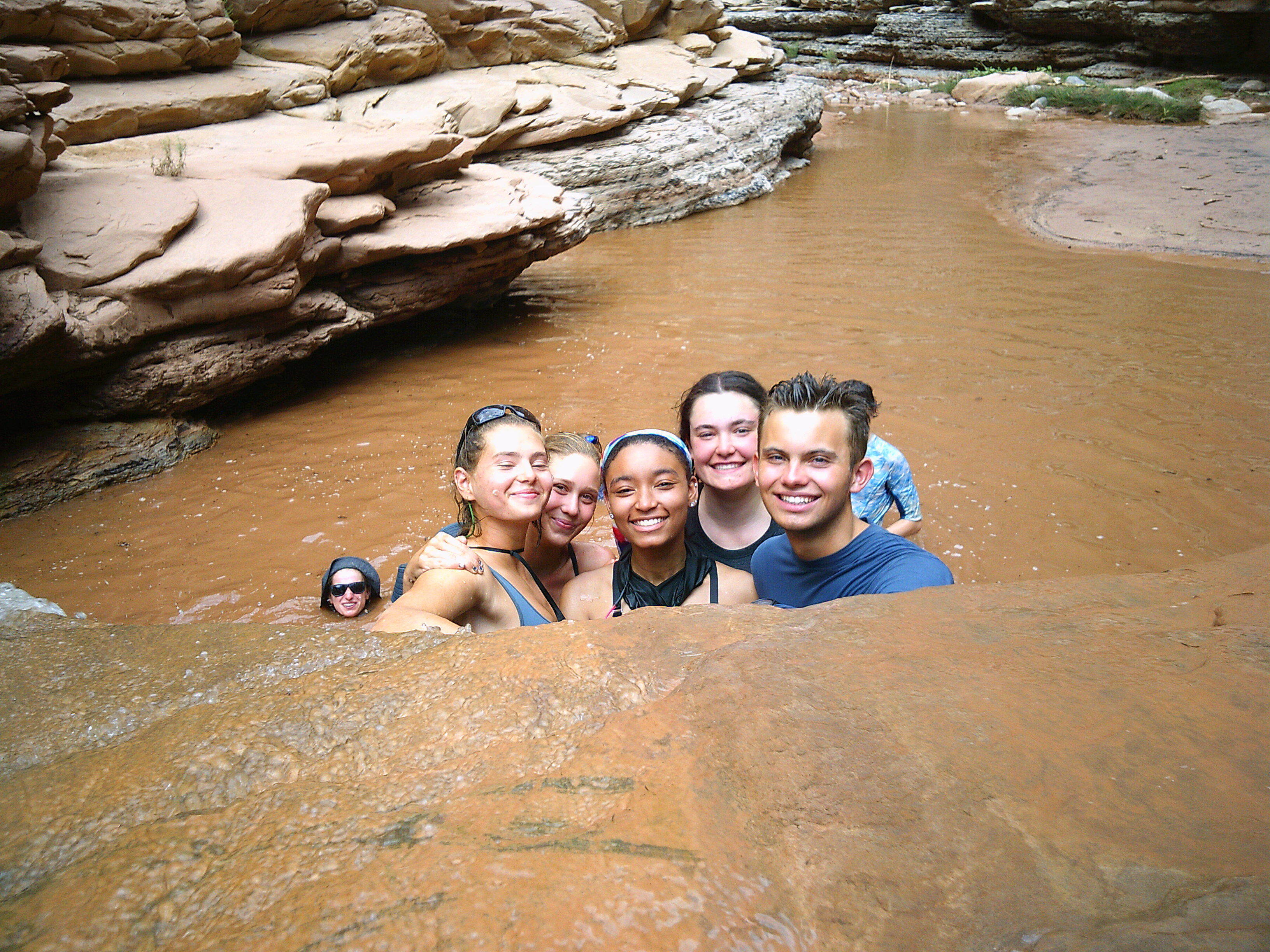 In a shallow, muddy river, a group of seven young people are gathered closely together, smiling at the camera. The water is a murky brown color, contrasting with the rocky, layered formations on the riverbank. The group appears to be enjoying a casual swim or wade in a natural setting, with some partially submerged in the water. The overall atmosphere is one of camaraderie and outdoor fun.