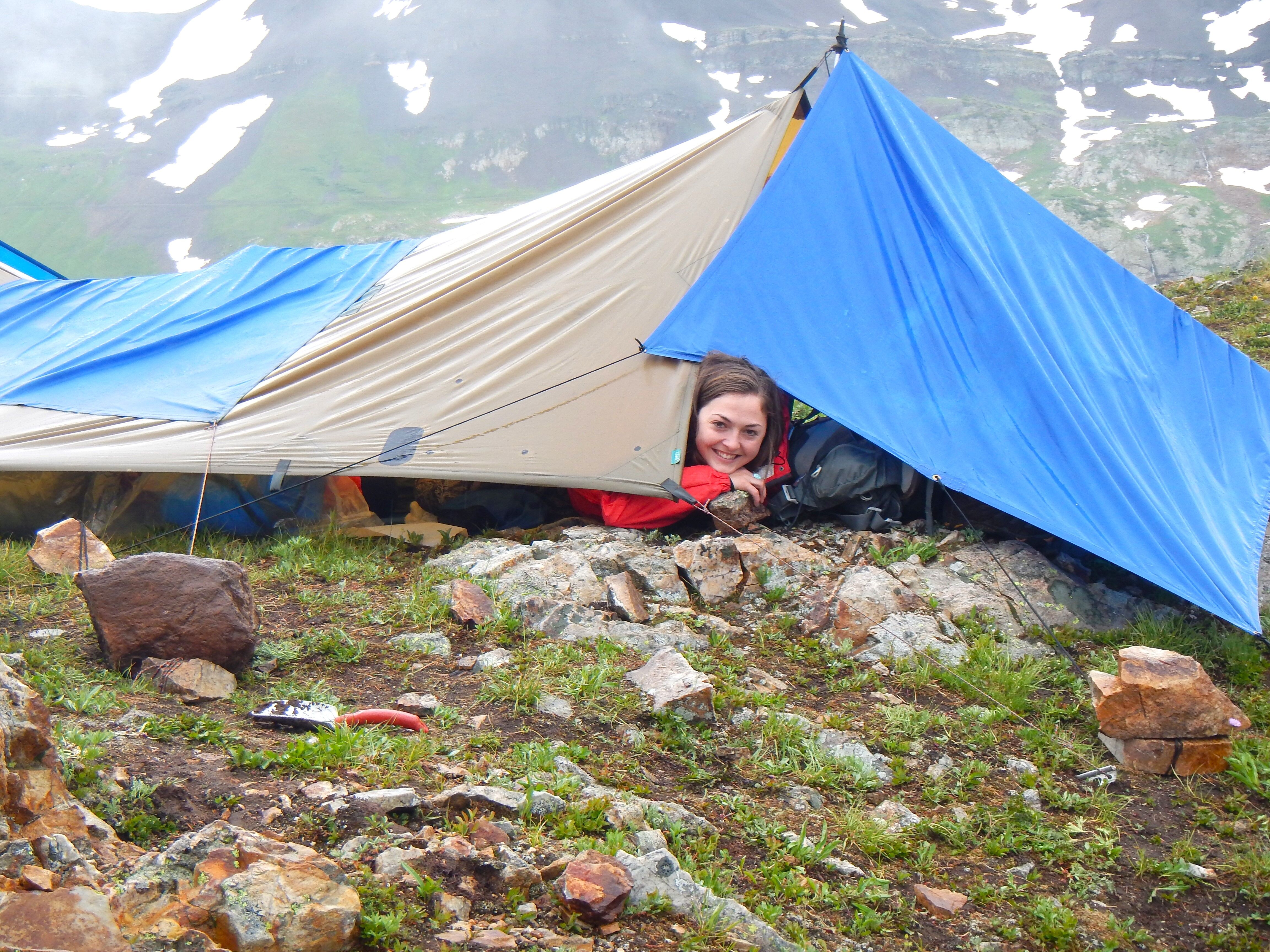 A woman is peeking out of a tent pitched on a rocky, grassy hillside. The tent is a mix of tan and blue fabric, blending with the natural surroundings. The landscape includes patches of green vegetation and scattered rocks, suggesting a mountainous or wilderness area. The weather appears overcast, with a hint of mist or fog in the background, adding to the remote and possibly challenging environment.