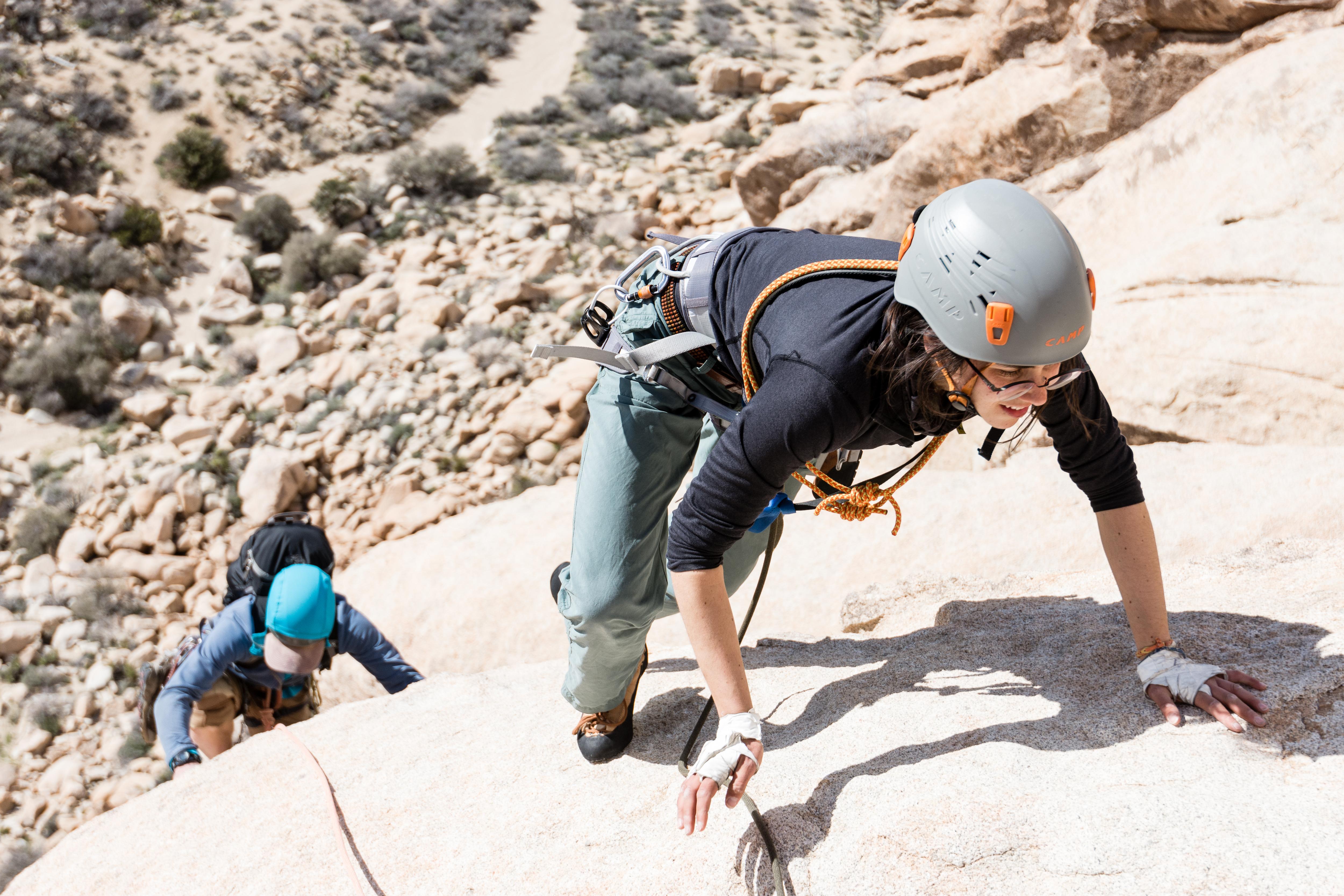 The image shows two people rock climbing on a sunny day. The person in the foreground is wearing a grey helmet, a black long-sleeved shirt, and light blue pants. They are using their hands to grip the rock face. The second person is further back and also climbing, wearing a blue hat. The rock is light-colored, and the background shows a rocky, desert-like landscape.